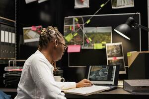 African american woman private detective studying clues in agency office. Police investigator sitting at desk, looking at suspect photo on laptop and reading criminal case information