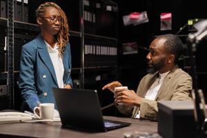 African american cops team working in archive to solve crime case. Man detective sitting at desk near shelf full of folders and files, discussing insight with woman partner at night time photo