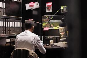 Police investigator working overtime analyzing evidence to solve crime. Criminalist sitting at desk, checking suspect photos on laptop and studying clues on detective board