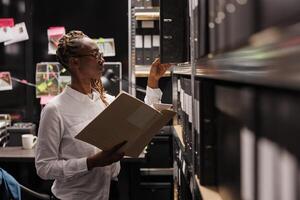 Police investigator studying archival crime case materials, working overtime in detective office room. African american policewoman searching evidence records and taking file from shelf photo