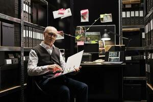 Elderly criminal investigator analyzing crime scene report, checking criminology files. Fbi agent working late at night at suspect profile in arhive room, planning confidential federal investigation photo