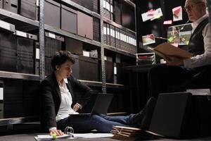 Private detective woman sitting on floor in arhive room, analyzing criminology report. Police officer working overtime at criminal case, checking crime scene evidence trying to catch suspect photo