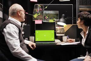 Police officers looking at laptop computer with greenscreen display, working late at night at criminal case in arhive room. Private detectives discussing crime scene evidence. Isolated template screen photo