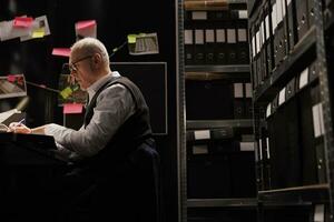 Elderly investigator working overtime at crime scene report, analyzing evidence files in arhive room. Overworked private detective checking criminology documents, planning strategy to catch suspect photo