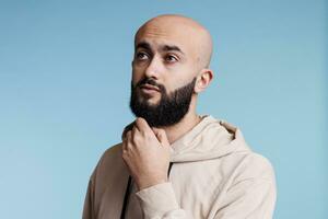 Arab man looking away with pensive facial expression and rubbing chin thoughtfully. Young bald bearded person thinking and making decision while posing with puzzled gesture photo