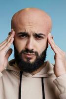 Arab man feeling pain in head while rubbing temples and looking at camera. Young handsome person suffering from headache with exhausted facial expression studio close up portrait photo