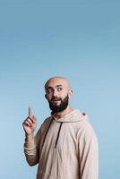 Inspired arab man pointing up while making imagination and inspiration gesture. Young handsome person raising index finger upwards and looking away while posing in studio photo