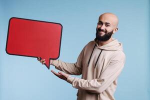 Cheerful arab man promoting new product with comic balloon red banner portrait. Smiling person holding empty dialog bubble and looking at camera with happy facial expression photo