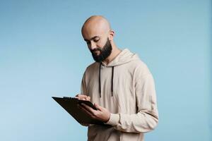 Concentrated arab man planning and writing checklist while holding clipboard. Young focused person wearing casual hoodie checking items in list and taking notes in studio photo