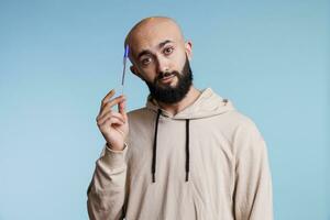 Thoughtful arab man holding pen and tilting head while looking at camera with pensive facial expression. Young bald bearded person planning, thinking and dreaming studio portrait photo