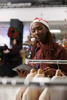 ventas gerente cheques inventario lista con ropa artículos en Al por menor almacenar, trabajando en valores durante Navidad fiesta estación. mujer con Papa Noel sombrero contando mercancías en bastidores, festivo boutique. foto