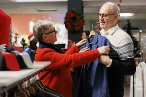 mayor Pareja examinando chaquetas americanas en bastidores en centro comercial ropa almacenar, mirando para formal atuendo a vestir en Navidad cena festividad. mayor clientes buscando para elegante ropa en festivo tienda. foto