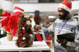 Selective focus of worker counting ties next to racks in clothing store, wearing santa hat during sales season. Young man managing stock inventory with list, arranging accessories in displayed boxes. photo