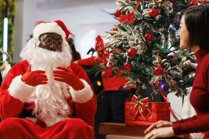 Mall worker Santa Claus sitting next to xmas tree with asian client in festive decorated retail store. Fashion boutique employee offering gift to happy customer, discussing about winter wishes. photo