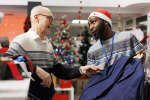 Male employee recommending clothes for senior customer, looking to buy formal outfit for christmas eve dinner. Elderly man asking worker for assistance to choose perfect fit clothing items. photo