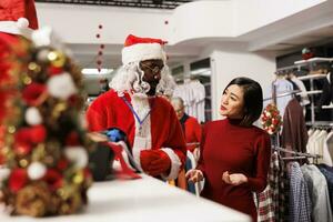 Store worker in santa suit helps client and presenting new clothing collection at shopping mall, talking to asian woman about christmas present ideas. Festive person in costume greeting buyer. photo