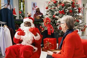 Santa claus employee gives presents to elderly woman in store, offering festive box next to xmas tree at mall. Senior person feeling cheerful after receiving goods for free, seasonal sales. photo