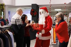 People finding out contest winner in shop after man dressed as santa drawing prize names from red box, making someone happy with free clothing items during christmas. Clients waiting impatient. photo