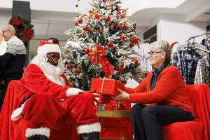 Senior woman sitting with santa claus in store, receiving beautiful christmas present from employee next to festive decorations. African american man spreading holiday spirit in shopping mall. photo