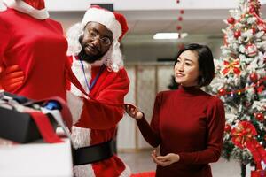 Retail worker showing items to buyer in clothing store, recommending clothes and accessories to wear on christmas eve dinner. Asian woman talking to assistant in festive santa suit. photo
