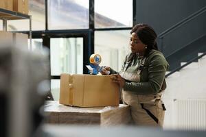 African american manager using adhesive tape in storehouse, working at customers orders. Storehouse manager preparing clients packages, checking products during storage room inventory photo