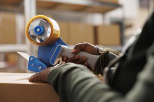 African american employee preparing customers orders, putting adhesive tape on cardboard box before shipping packages. Stockroom supervisor working at goods inventory in storage rooom. Close up photo