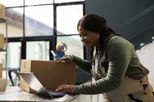 Storehouse supervisor checking online orders on laptop computer, analyzing delivery details while preparing packages. African american employee analyzing merchandise logistics in storage room photo