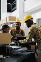 Postal service workers checking parcel invoice on laptop and clipboard. African american men and woman storehouse employees team managing inventory documentation at reception photo