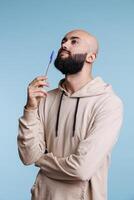 Thoughtful arab man holding pen and looking upwards in contemplation. Pensive young adult bald bearded person standing in thinking pose with questioned unsure facial expression photo