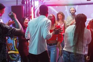 Friends dancing at disco party and clubbing while enjoying nightlife leisure activity. Diverse people making moves to electronic music rhythm on dancefloor in nightclub with spotlights photo