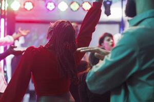 africano americano mujer en rojo blusa saltando y bailando en concurrido discoteca. joven clubbers teniendo divertido y ir de fiesta en pista de baile con focos a discoteca en oscuro club foto