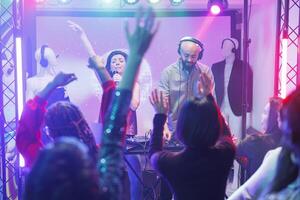 Crowd dancing and raising hands at band concert in nightclub. Singer and dj performing on stage with spotlights and engaging diverse people partying at discotheque on dancefloor photo