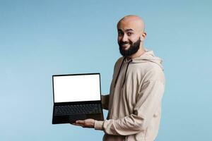 Cheerful smiling arab man advertising laptop with white empty screen mock up portrait. Happy person showing portable computer with blank display for software promotion and looking at camera photo