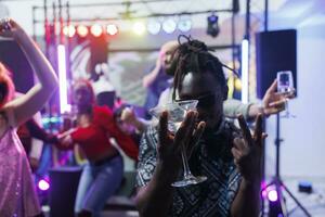 African american clubber drinking alcohol and partying in nightclub. Young cheerful man holding glass with beverage and showing gestures at camera while having fun at discotheque photo