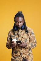 Positive man searching information on internet, scrolling on social media in studio over yellow background. African american young adult drinking coffee while typing message on mobile phone photo