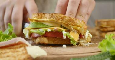 vrouw chef bezuinigingen club belegd broodje in een voor de helft door scherp mes Aan een houten bord Bij de keuken. groot smakelijk belegd broodje met ham, tomaat plakjes en stukken van kaas, sla en mayonaise saus. ultra 4k video