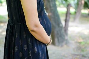 un embarazada indio dama poses para al aire libre el embarazo disparar y manos en barriga, indio embarazada mujer pone su mano en su estómago con un maternidad vestir a sociedad parque, embarazada fuera de maternidad disparar foto