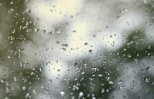 A photo of rain drops on the window glass with a blurred view of the blossoming green trees. Abstract image showing cloudy and rainy weather conditions