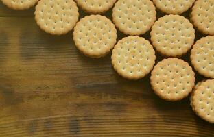 A round sandwich cookie with coconut filling lies in large quantities on a brown wooden surface. Photo of edible treats on a wooden background with copy space