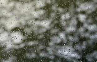 A photo of rain drops on the window glass with a blurred view of the blossoming green trees. Abstract image showing cloudy and rainy weather conditions