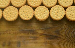 A round sandwich cookie with coconut filling lies in large quantities on a brown wooden surface. Photo of edible treats on a wooden background with copy space