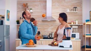 Man joggle with oranges for wife in kitchen while she is preparing fruits for smoothie. Cheerful family making together organic healthy fresh nutritious tasty juice for breakfast from fresh fruits while on a diet. photo