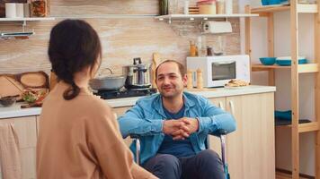 Portrait of happy disabled man in wheelchair looking at wife in kitchen. Guy with paralysis handicap disability handicapped difficulties getting help for mobility from love and relationship photo