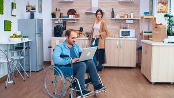 Handicapped entrepreneur in wheelchair working on laptop while wife is preparing meal in kitchen. Man with paralysis handicap disability handicapped difficulties getting help for mobility from love and relationship photo