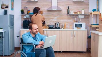 Disabled man waving on video conference using laptop in kitchen. Corporate man with paralysis handicap disability handicapped difficulties working after accident having internet online video call conference communication for professional use. photo