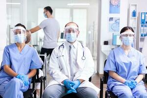 Group of medical personnel in hospital waiting area with visor and face mask against covid-19 to prevent infection. Wearing white coat and stethoscope. photo