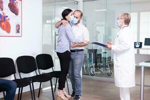 Disappointed young couple in hospital waiting area from doctor unfavorable bad news during coronavirus outbreak. Medic wearing face mask against infection with covid-19. photo