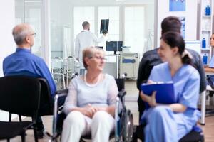 Nurse filing documents while talking with disabled senior woman in hospital waiting area. Patient asking for direction at hospital reception. photo