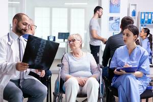 Doctor with stethoscope holding disabled senior woman radiography in wheelchair while talking with her in hospital waiting area. Patient asking about his appointment at clinic reception. photo