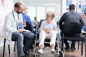 Doctor holding tablet pc with x-ray while explaining diagnosis to disabled elderly woman in wheelchair. Handicapped patient in hospital waiting area. Man in examination room. photo